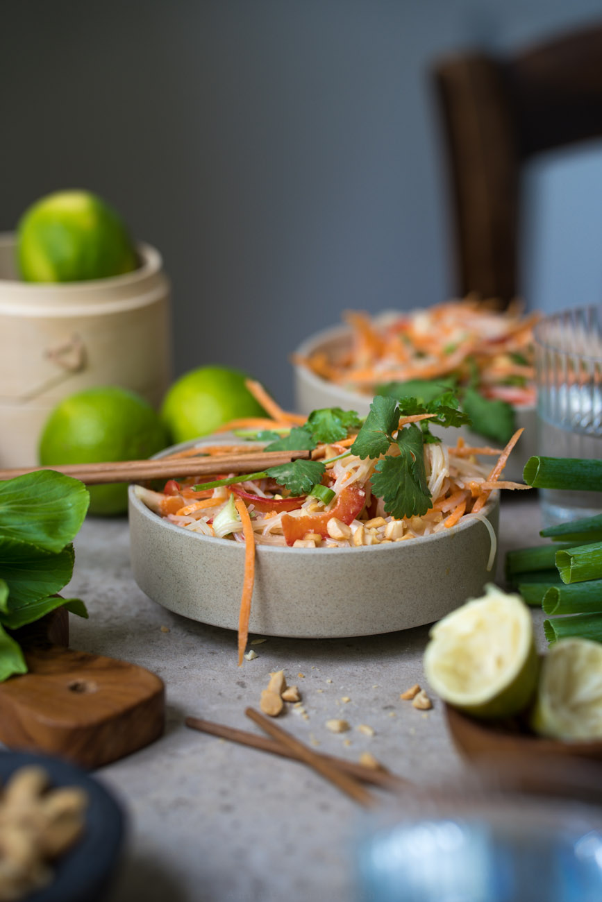 Glasnudelsalat mit Erdnusssoße in einer beigen Keramikschale auf einem Tisch. Im Hintergrund steht eine weitere Schale. Sonst sieht man ausgepresste Limetten im Vordergrund, Pak Choi am Rand, Wassergläser auf dem Tisch.