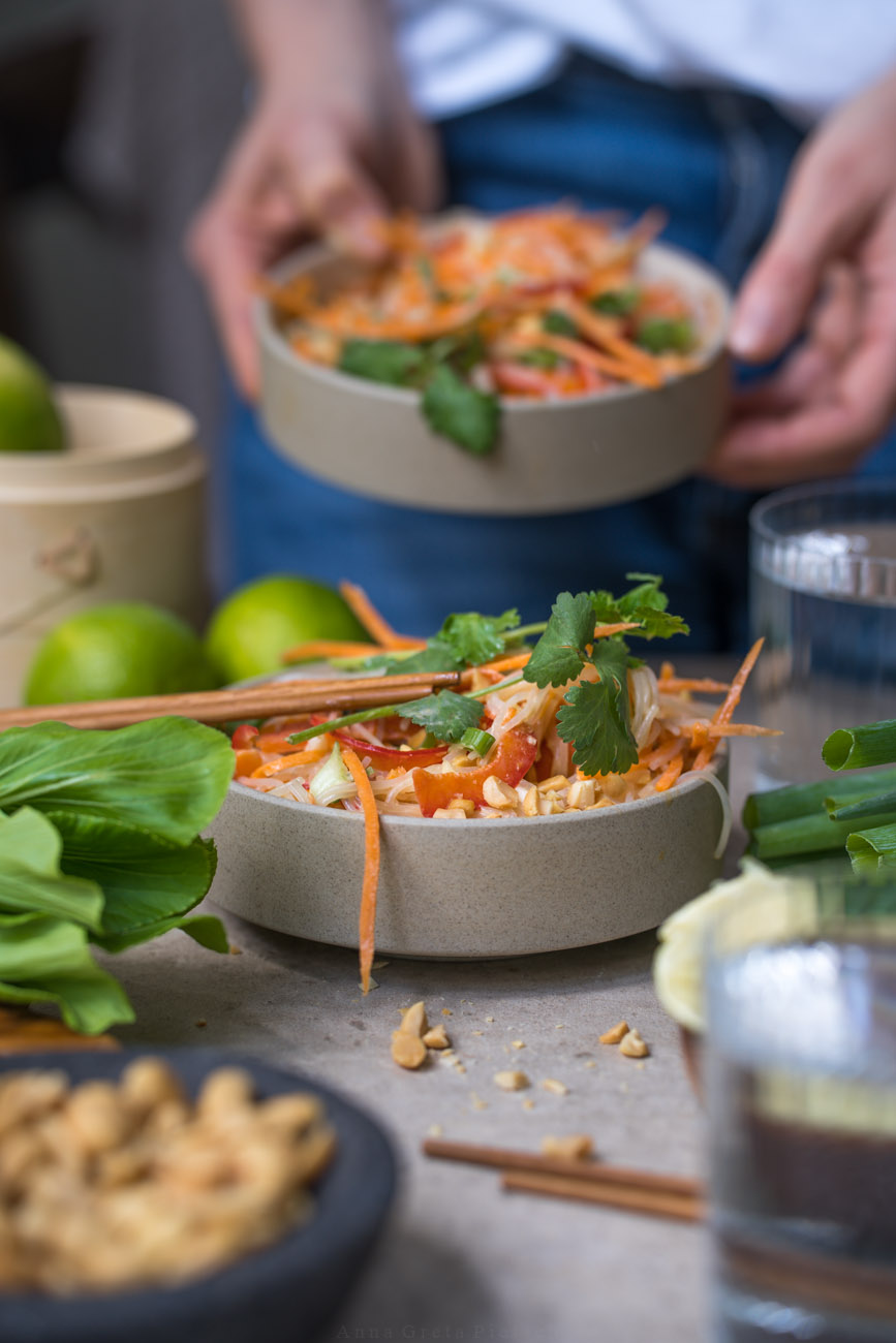 Glasnudelsalat mit Erdnusssoße in einer beigen Keramikschale auf einem Tisch. Im Hintergrund hält eine Person eine weitere gefüllte Schale. Sonst sieht man ausgepresste Limetten im Vordergrund, Pak Choi am Rand, Wassergläser auf dem Tisch.