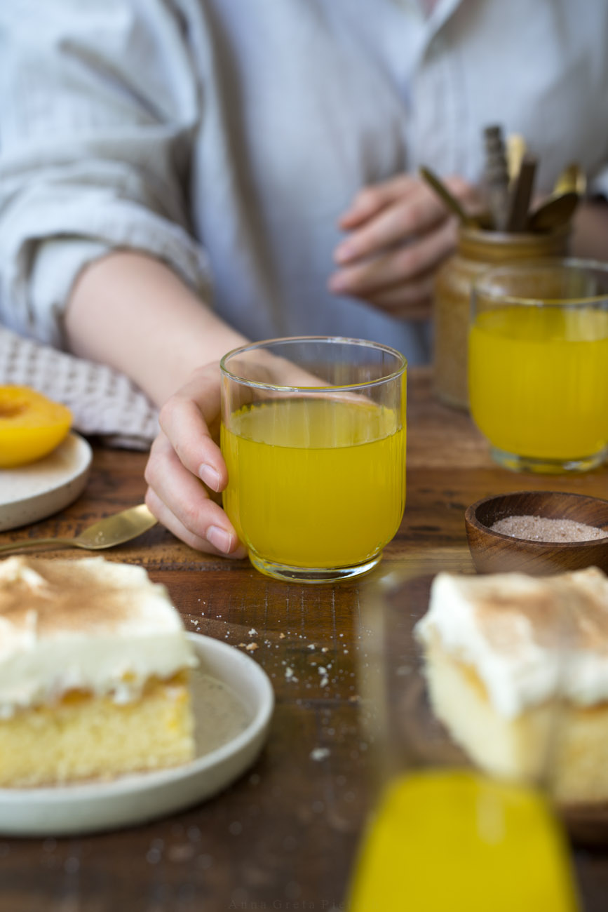 Ein Glas mit Orangenlimonade auf einer Kaffeetafel, gehalten von einer Hand. Die Person sitzt dahinter am Tisch.