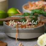 Glasnudelsalat mit Erdnusssoße in einer beigen Keramikschale auf einem Tisch. Im Hintergrund steht eine weitere Schale. Sonst sieht man ausgepresste Limetten im Vordergrund, Pak Choi am Rand, Wassergläser auf dem Tisch.