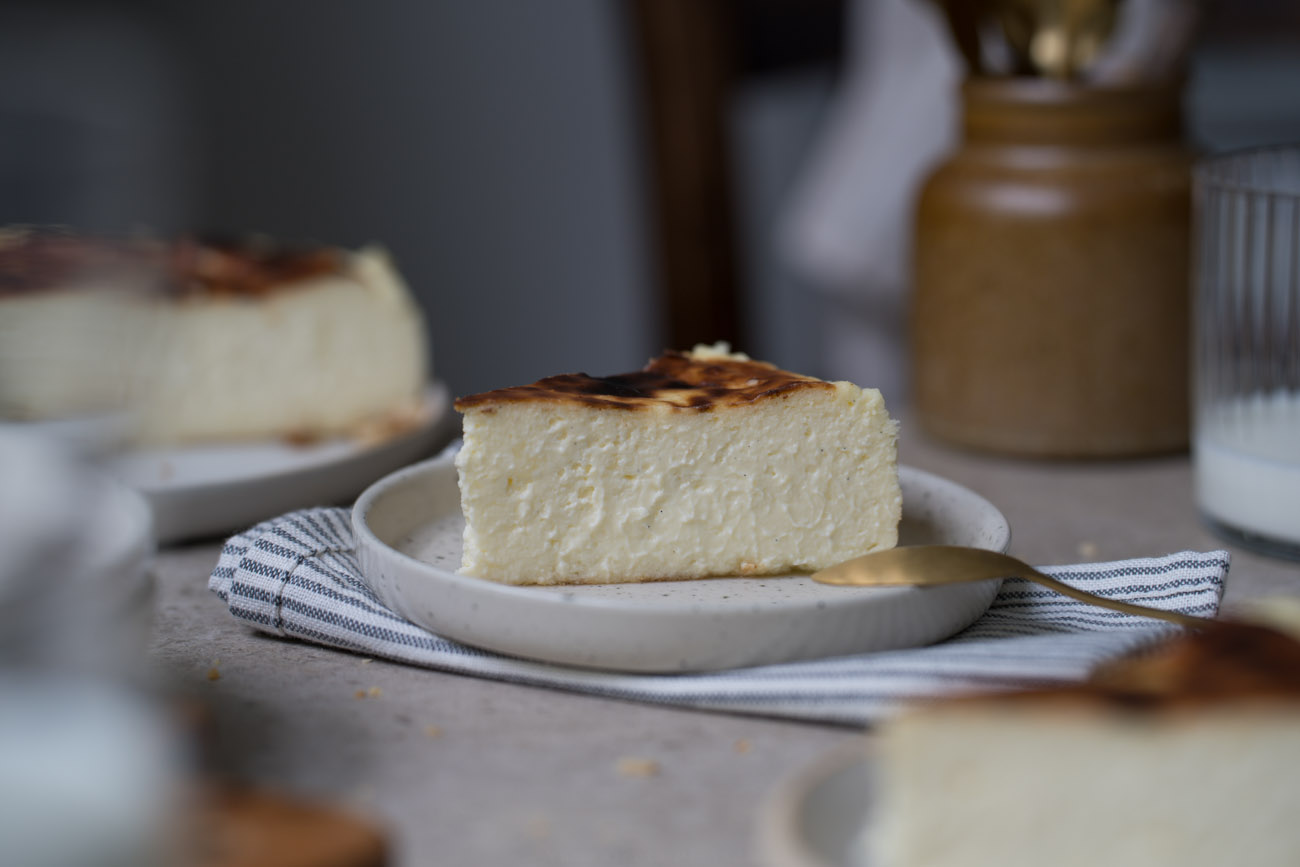 Ein Stück San Sebastian Cheesecake auf einem kleinen Teller steht in der Mitte einer Kaffeetafel. Man sieht im Hintergrund und Vordergrund noch weitere Kuchenstücke, den Kuchen auf einer Kuchenplatte.