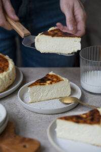 Ein Stück San Sebastian Cheesecake auf einem kleinen Teller steht in der Mitte einer Kaffeetafel. Man sieht im Hintergrund und Vordergrund noch weitere Kuchenstücke, den Kuchen auf einer Kuchenplatte. Eine Person hält im Hintergrund einen Tortenheber mit einem Stück des Kuchens darauf.