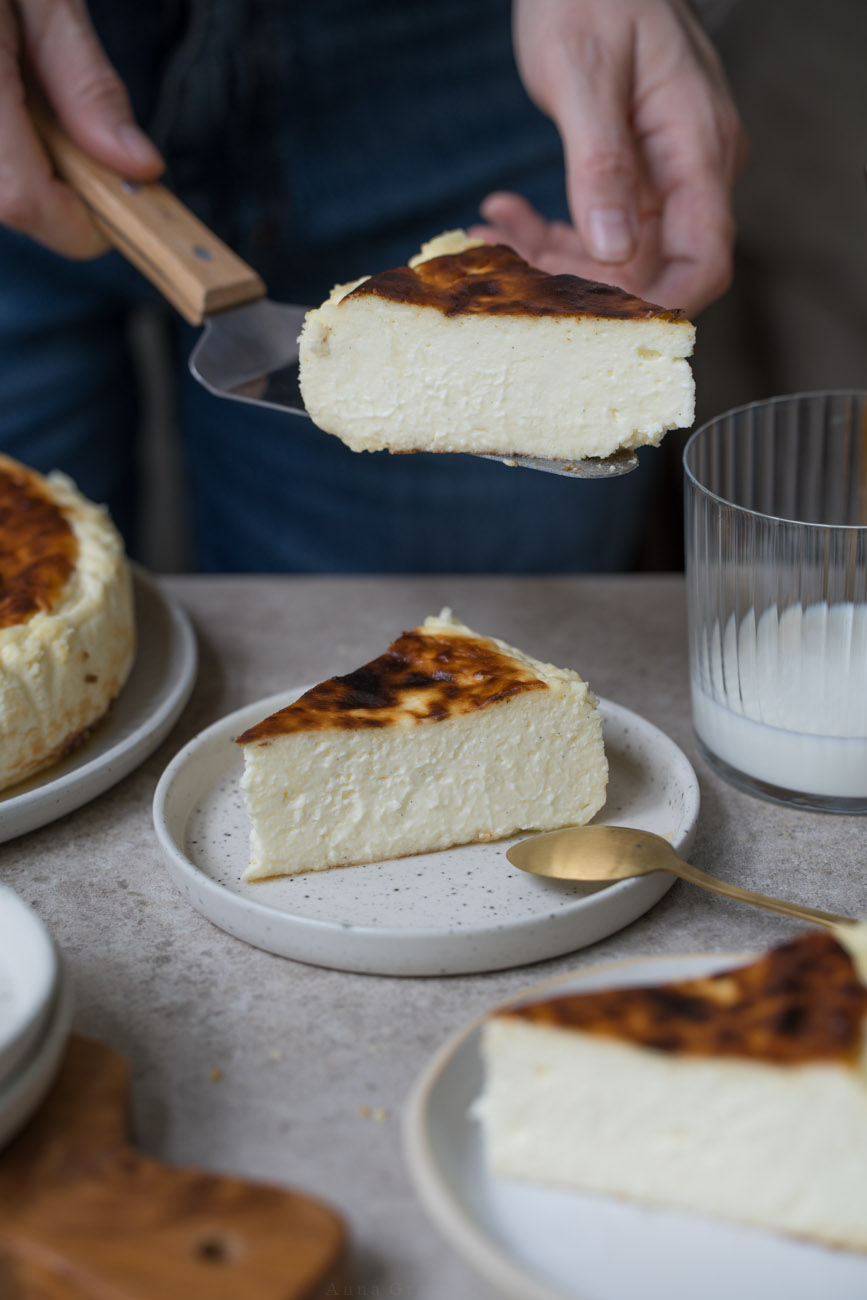 Ein Stück San Sebastian Cheesecake auf einem kleinen Teller steht in der Mitte einer Kaffeetafel. Man sieht im Hintergrund und Vordergrund noch weitere Kuchenstücke, den Kuchen auf einer Kuchenplatte. Eine Person hält im Hintergrund einen Tortenheber mit einem Stück des Kuchens darauf.