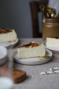 Ein Stück San Sebastian Cheesecake auf einem kleinen Teller steht in der Mitte einer Kaffeetafel. Man sieht im Hintergrund und Vordergrund noch weitere Kuchenstücke, den Kuchen auf einer Kuchenplatte.