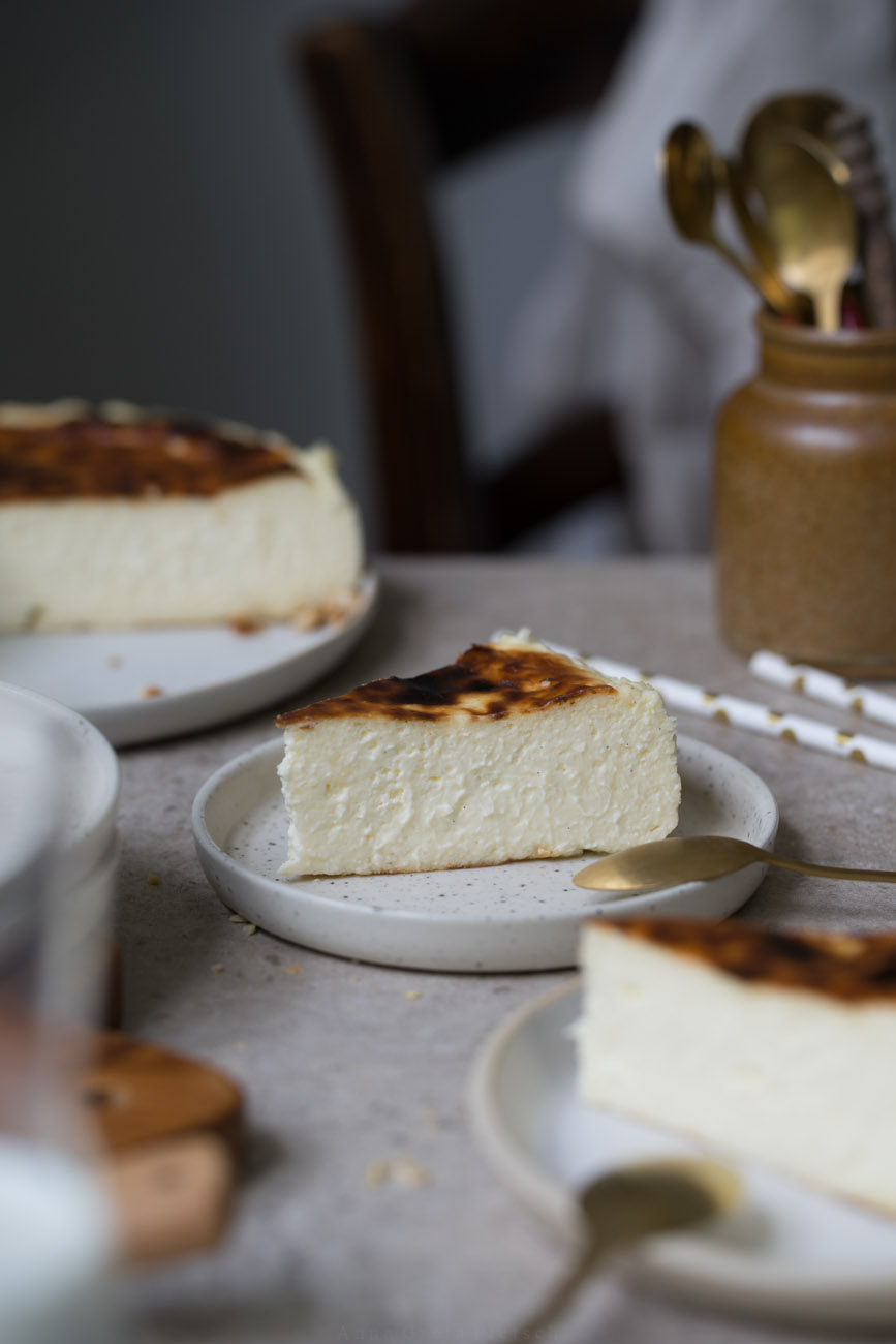 Ein Stück San Sebastian Cheesecake auf einem kleinen Teller steht in der Mitte einer Kaffeetafel. Man sieht im Hintergrund und Vordergrund noch weitere Kuchenstücke, den Kuchen auf einer Kuchenplatte.