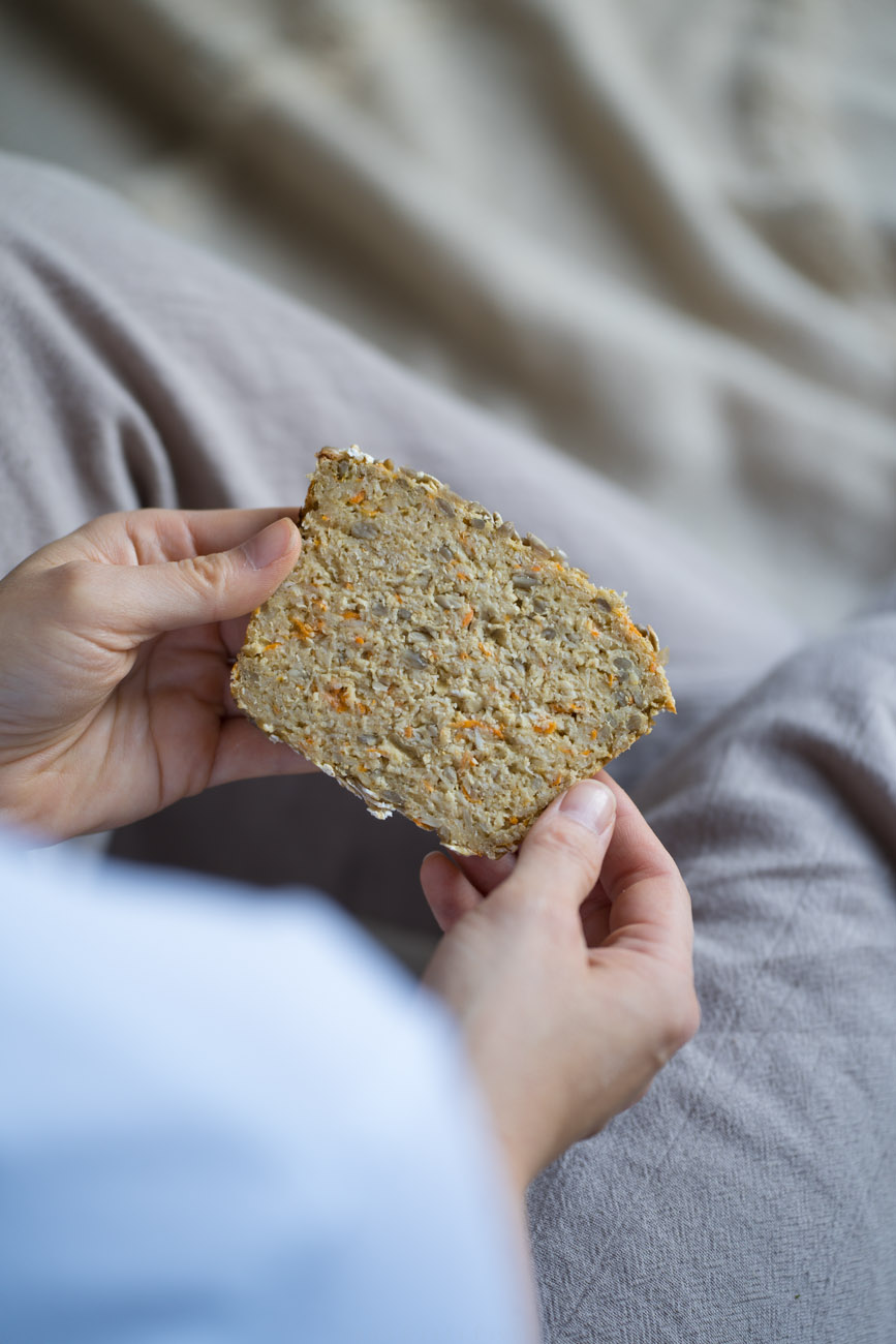 Eine PErson sitzt aif einer Decke und hält eine Scheibe Eiweißbrot mit Saaten in den Händen. Das Foto wurde über die Schulter fotografiert.