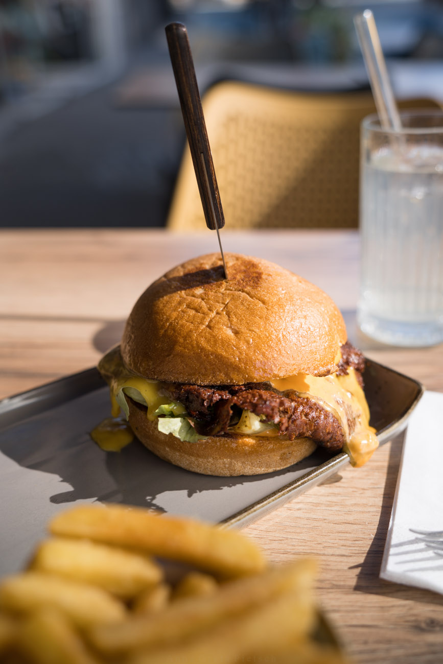 Ein glutenfreier Burger auf einem Tisch/Teller. Im Hintergrund steht ein Getränk und im Vordergrund sieht man Pommes.