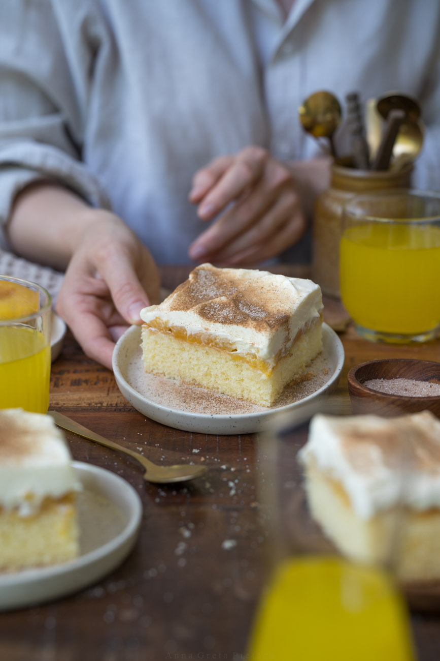 Eine Person mit einer beigen Bluse hält einen kleinen Kuchenteller mit Fantakuchen in einer Hand und stellt den Teller auf einer Kuchentafel ab.