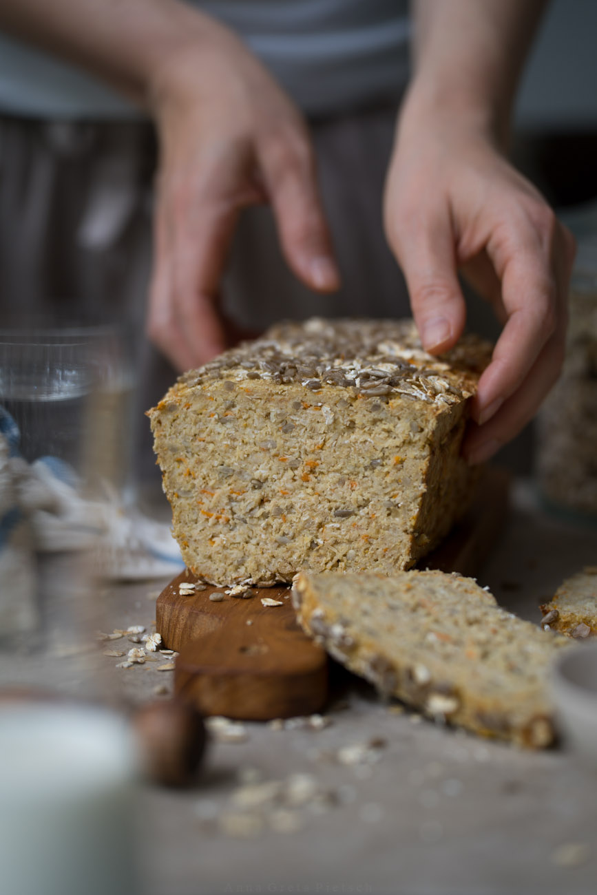 Ein glutenfreies Eiweißbrot mit Saaten steht auf einem Hozbrett auf einer hellen Tischplatte. Eine PErson hält es fest.