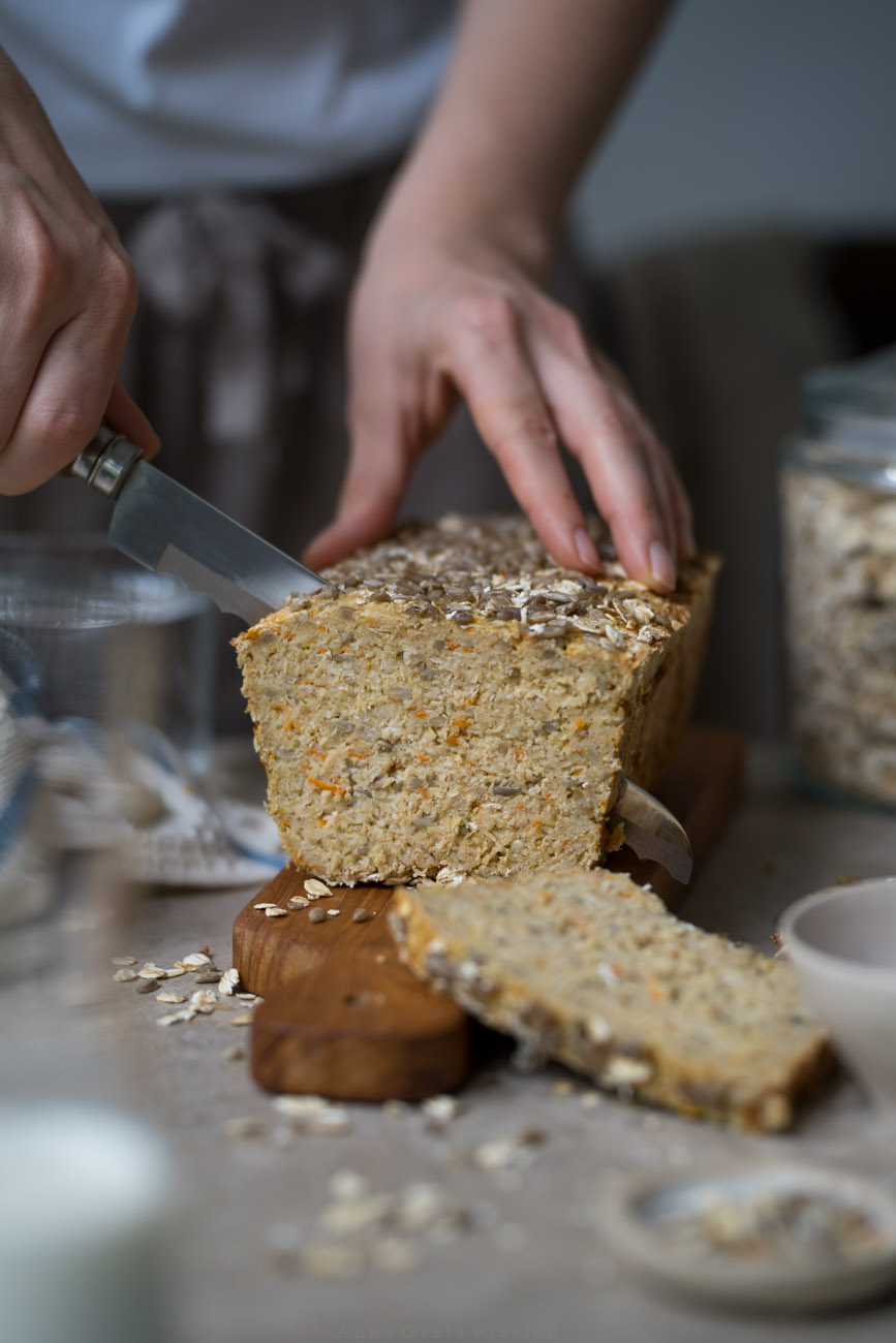 Ein glutenfreies Eiweißbrot mit Saaten steht auf einem Hozbrett auf einer hellen Tischplatte. Eine Person schneidet es mit einem Brotmesser an.