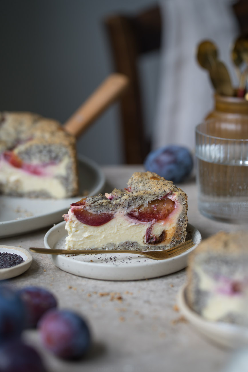 Ein Stück Käsekuchen mit Zwetschgen und Mohn steht auf einem kleinen Teller. Er steht in der Mitte einer Kaffeetafel. Man sieht im Hintergrund und Vordergrund noch weitere Kuchenstücke, den Kuchen auf einer Kuchenplatte.