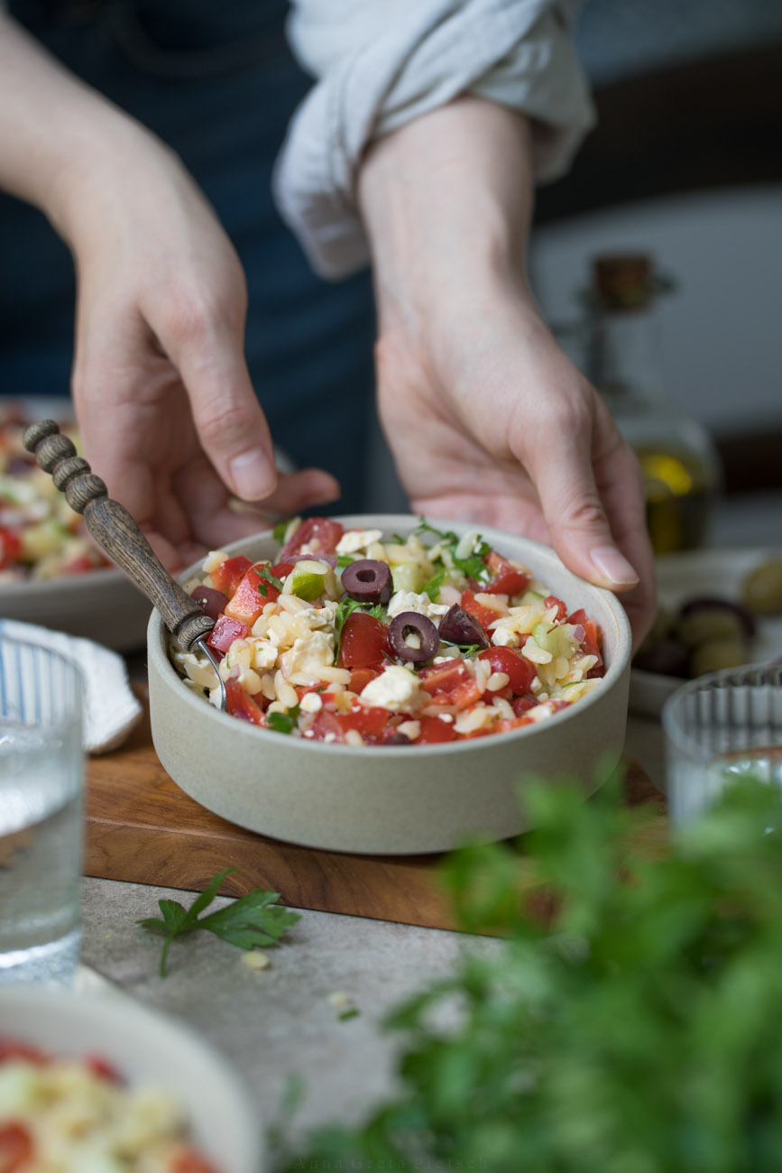 Ein gedeckter Tisch. Im Vordergrund steht PEtersilie, es stehen Wassergläser auf dem Tisch. In der Mitte stellt eine Person gerade eine Schale mit Orzo-Salat ab. Man erkennt die kleinen Nudeln, Paprika, Tomate, Oliven, Feta darin.