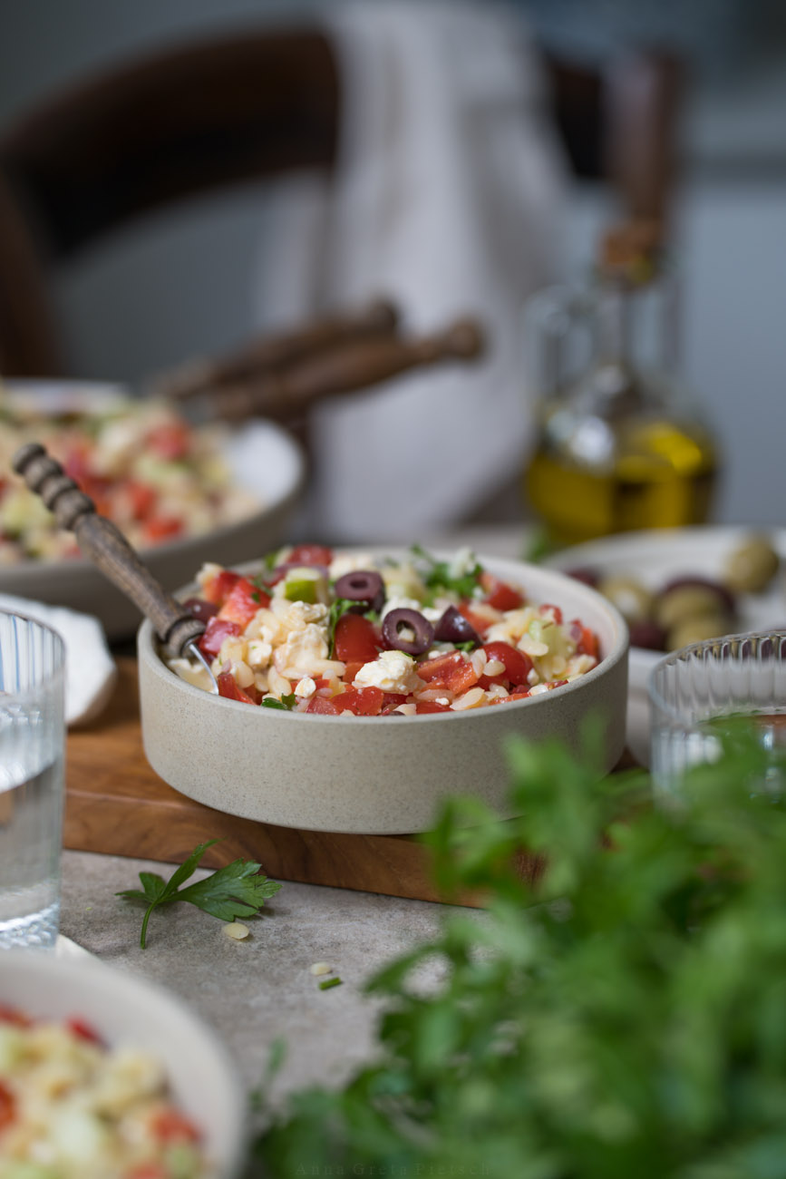 Ein gedeckter Tisch. Im Vordergrund steht PEtersilie, es stehen Wassergläser auf dem Tisch. In der Mitte steht ein Schälchen mit Orzo-Salat. Man erkennt die kleinen Nudeln, Paprika, Tomate, Oliven, Feta darin.