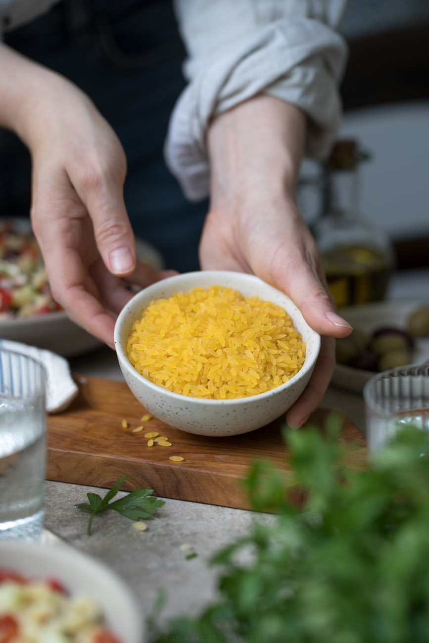 Eine Person stellt eine kleine Keramikschale mit rohen Orzo-Nudeln auf einem gedeckten Tisch ab.