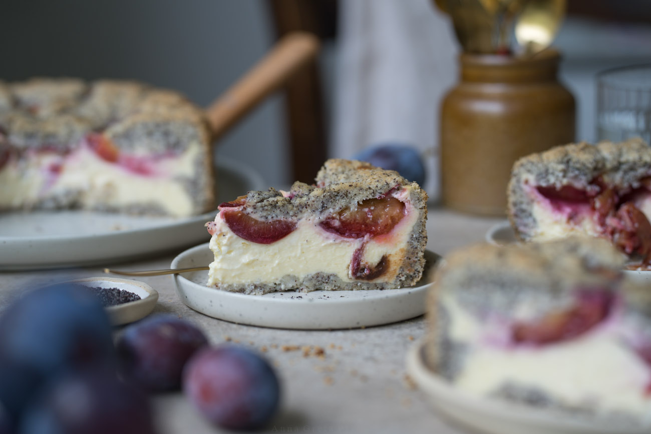 Ein Stück Käsekuchen mit Zwetschgen und Mohn steht auf einem kleinen Teller. Er steht in der Mitte einer Kaffeetafel. Man sieht im Hintergrund und Vordergrund noch weitere Kuchenstücke, den Kuchen auf einer Kuchenplatte.