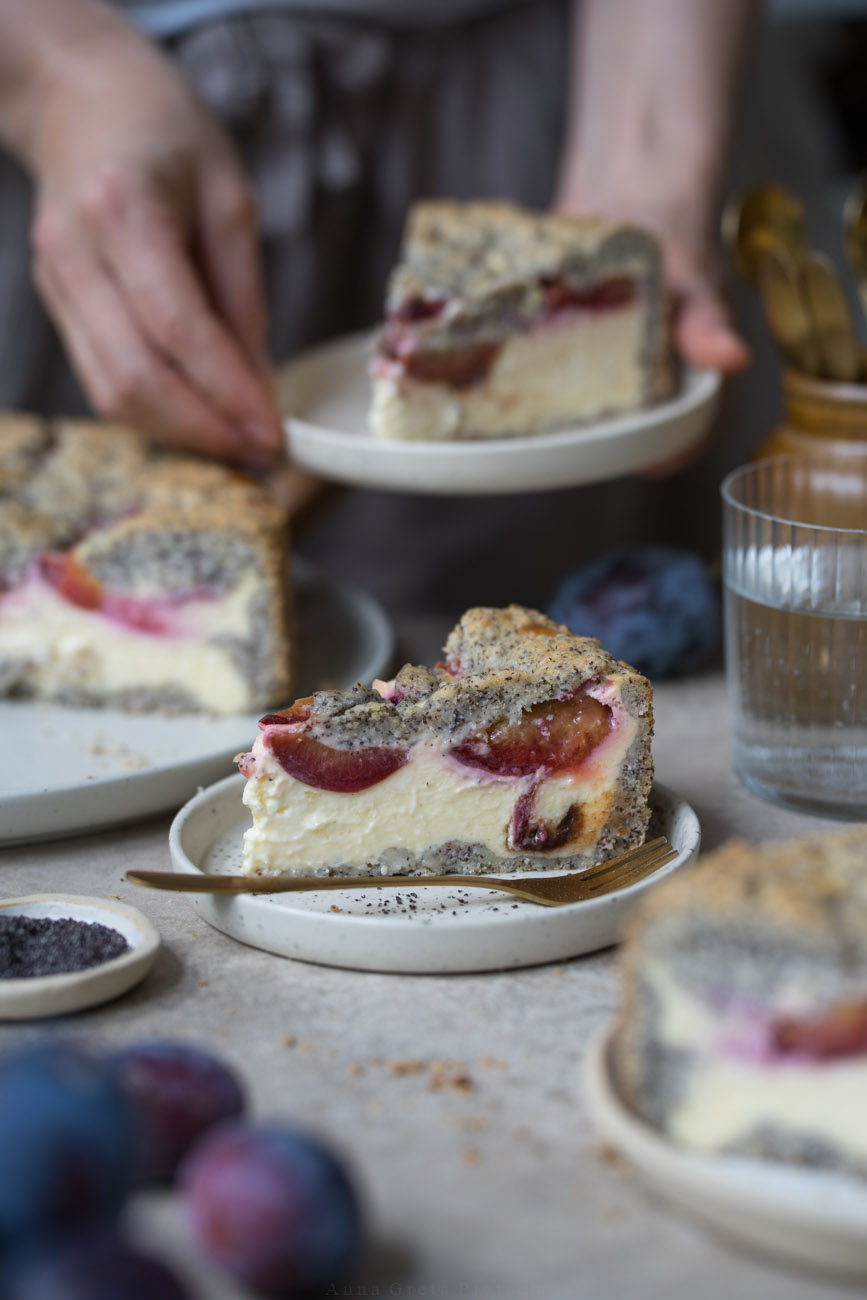 Ein Stück Käsekuchen mit Zwetschgen und Mohn steht auf einem kleinen Teller. Er steht in der Mitte einer Kaffeetafel. Man sieht im Hintergrund und Vordergrund noch weitere Kuchenstücke, den Kuchen auf einer Kuchenplatte. Im Hintergrund hält eine Person ein weiteres Kuchenstück auf einem Teller.