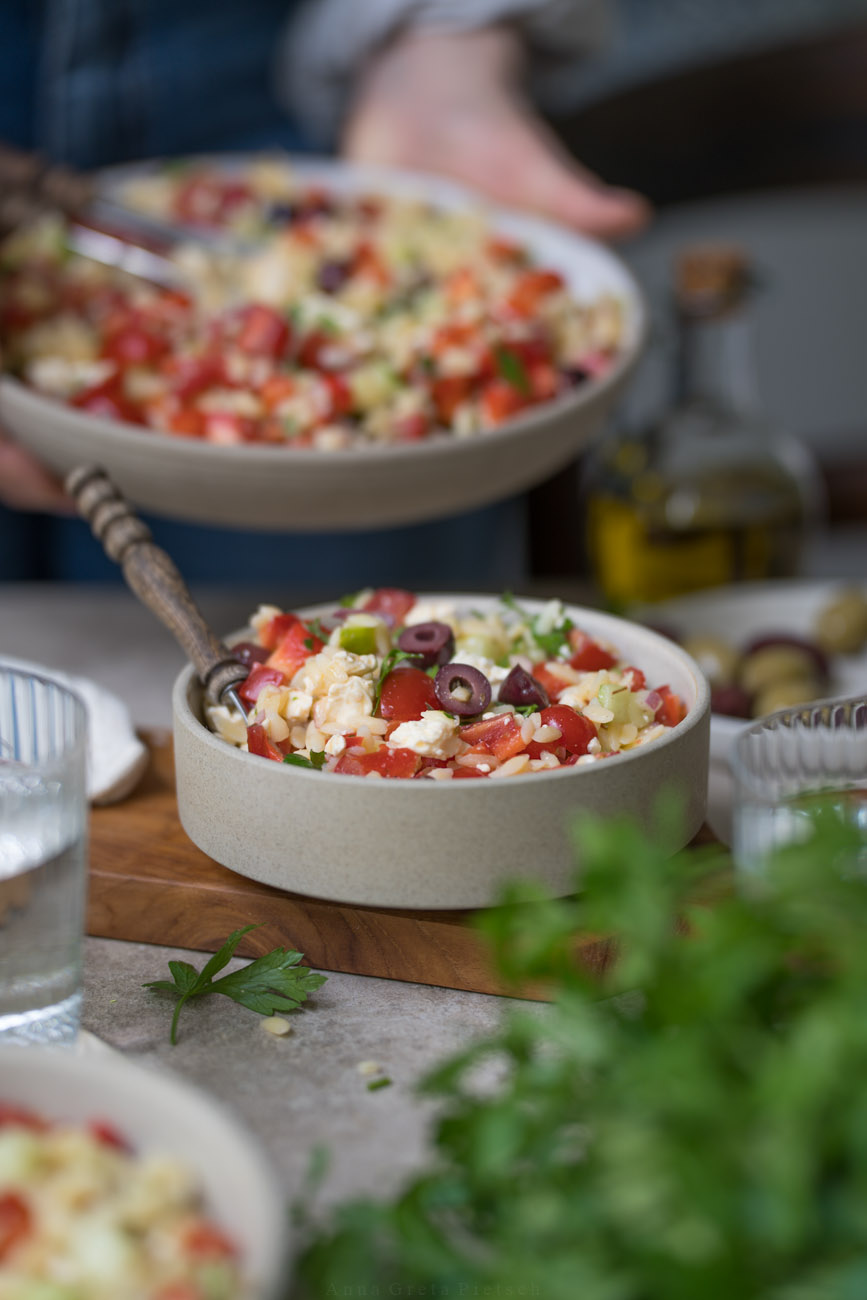 Ein gedeckter Tisch. Im Vordergrund steht PEtersilie, es stehen Wassergläser auf dem Tisch. In der Mitte steht ein Schälchen mit Orzo-Salat. Man erkennt die kleinen Nudeln, Paprika, Tomate, Oliven, Feta darin. Im Hintergrund hält eine Person noch eine größere Schale davon.