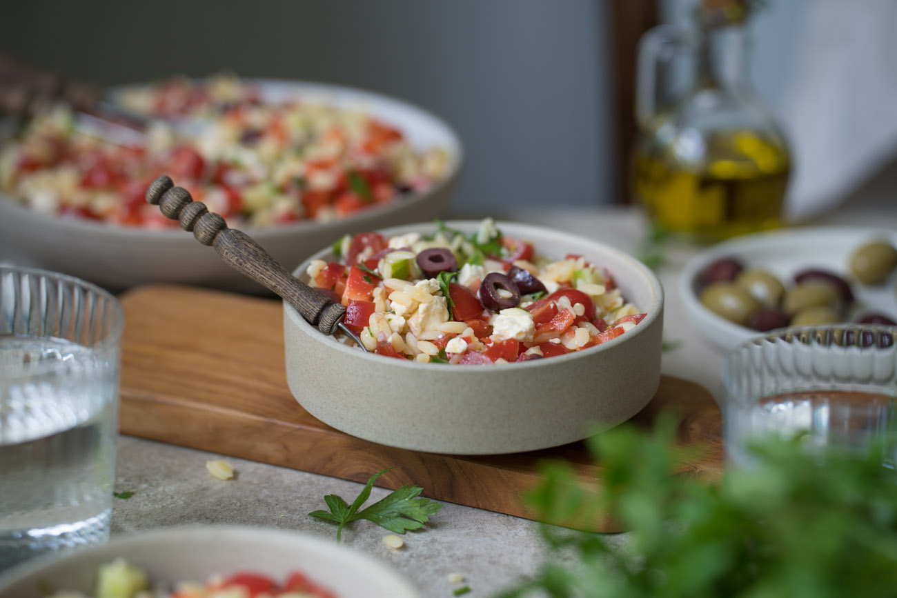 Ein gedeckter Tisch. Im Vordergrund steht PEtersilie, es stehen Wassergläser auf dem Tisch. In der Mitte steht ein Schälchen mit Orzo-Salat. Man erkennt die kleinen Nudeln, Paprika, Tomate, Oliven, Feta darin.