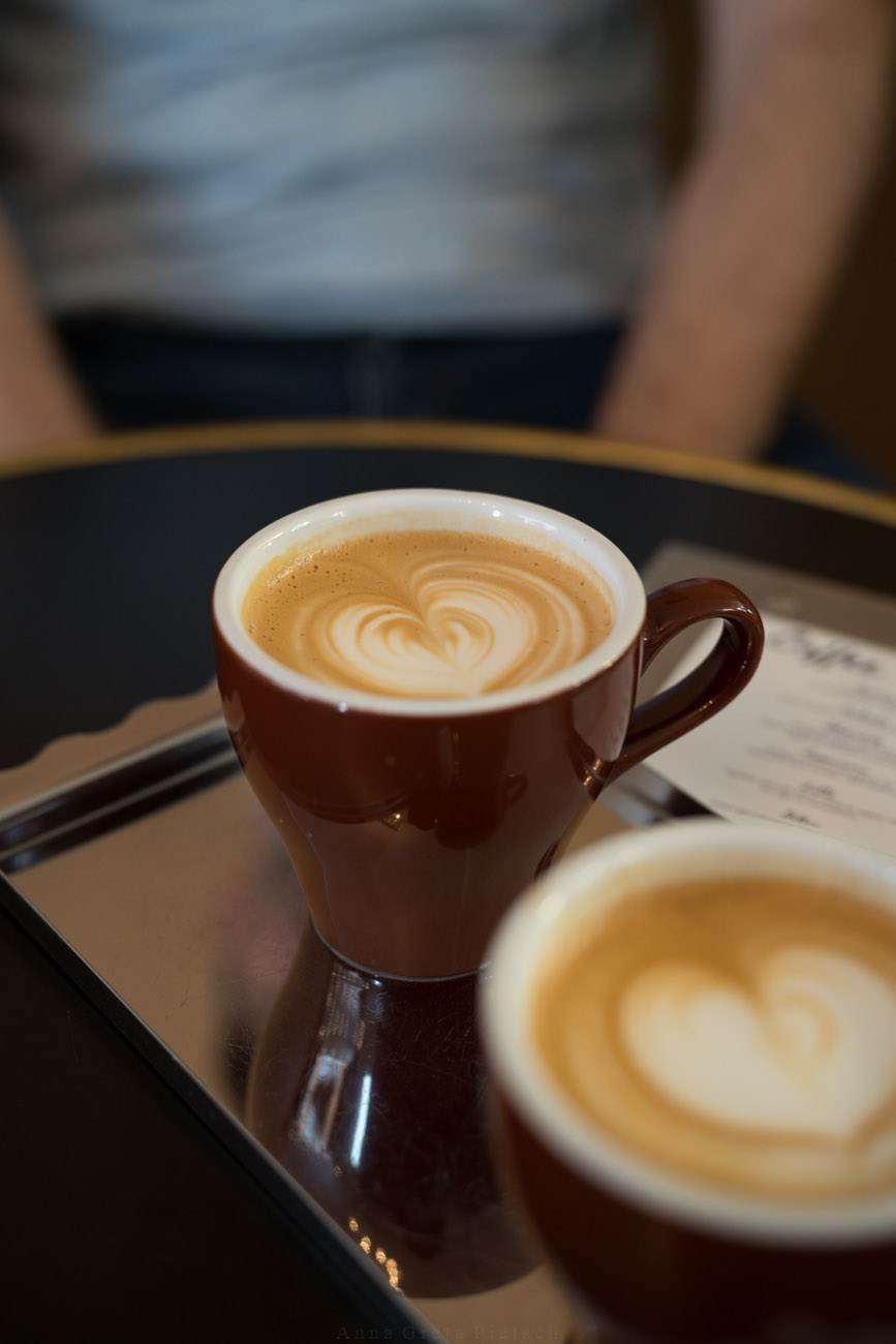 Zwei dunkelbraune Kaffeetassen auf einem Tisch. Gefüllt sind sie mit Cappuccino.