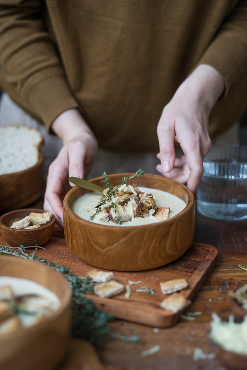 Man sieht einen Esstisch aus dunklem Holz, Darauf steht eine Holzschüssel mit einer hellen Suppe. Drumherum stehen weitere Schälen mit Suppe und Brot.
