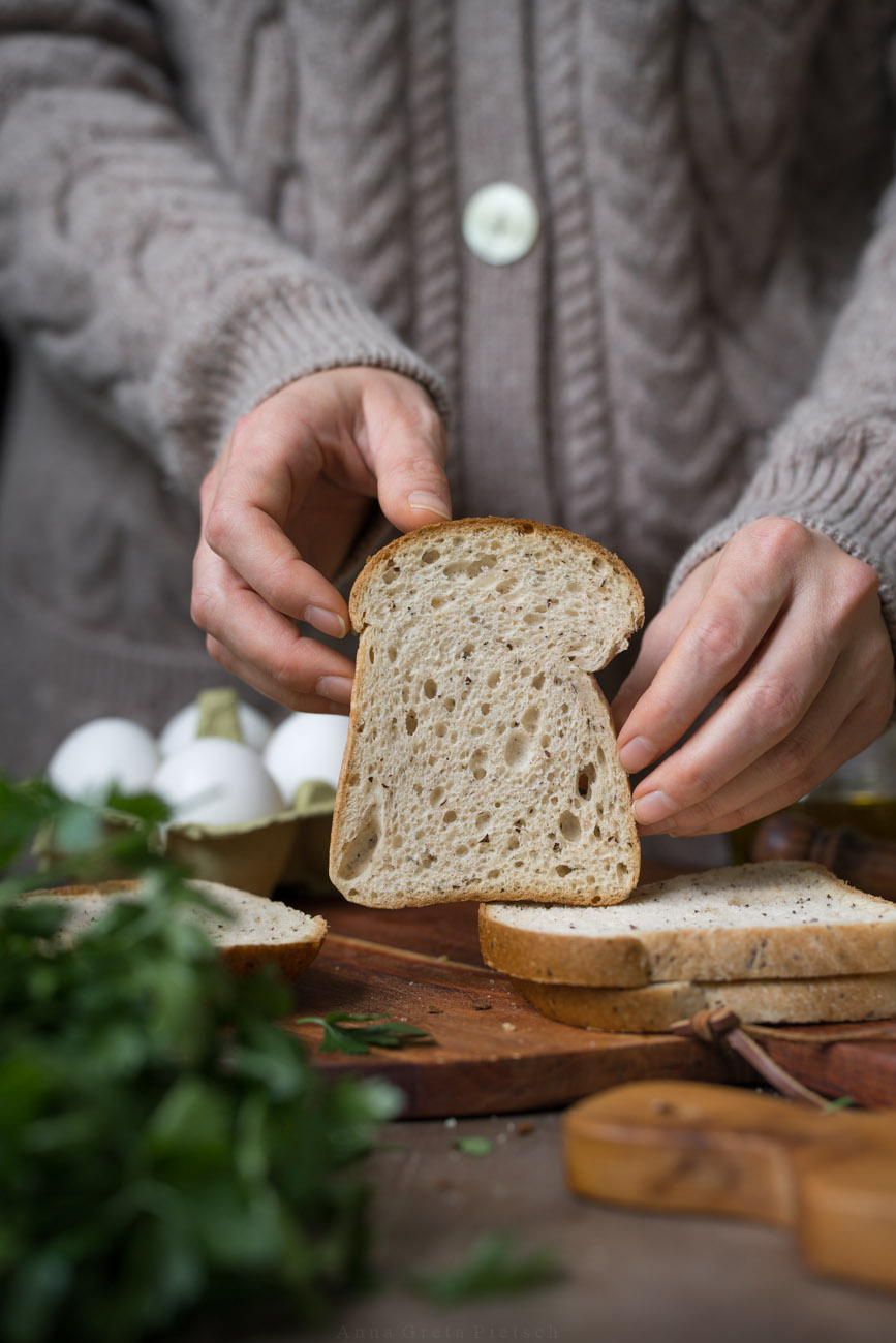 Eine Person mit einem dicken Strickpullover hält eine Brotscheibe auf einer Anrichte. Im Hintergrund steht man Eier und noch weitere Brotscheiben.