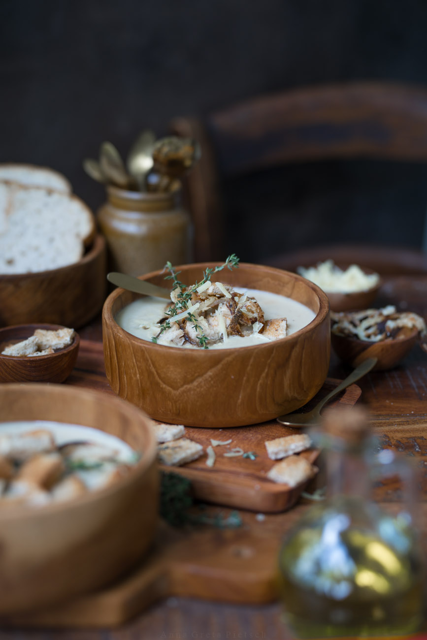 Man sieht einen Esstisch aus dunklem Holz, Darauf steht eine Holzschüssel mit einer hellen Suppe. Drumherum stehen weitere Schälen mit Suppe und Brot.