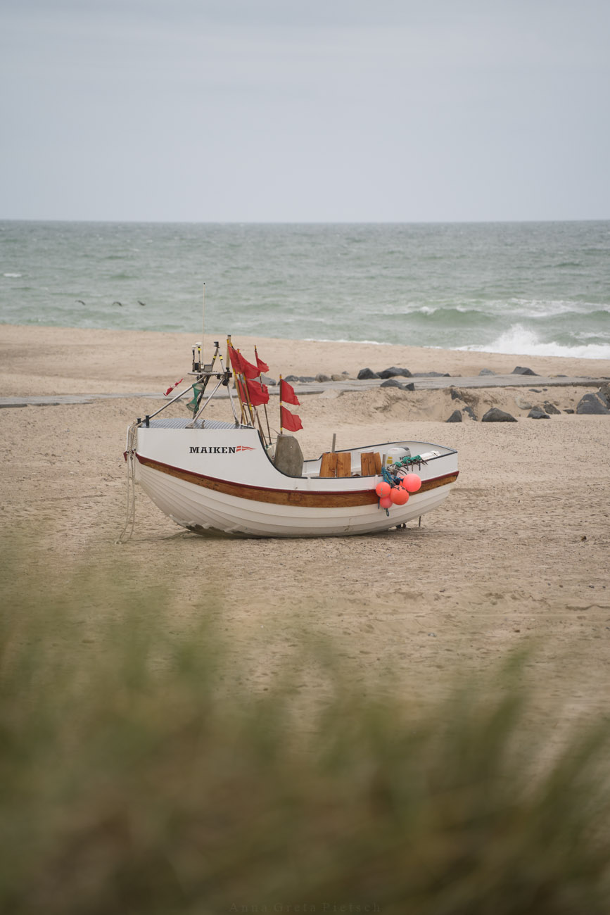 Ein kleines Fischerboot liegt in Dänemark auf dem Sand am Strand. Im Vordergrund sieht man Dünengras.