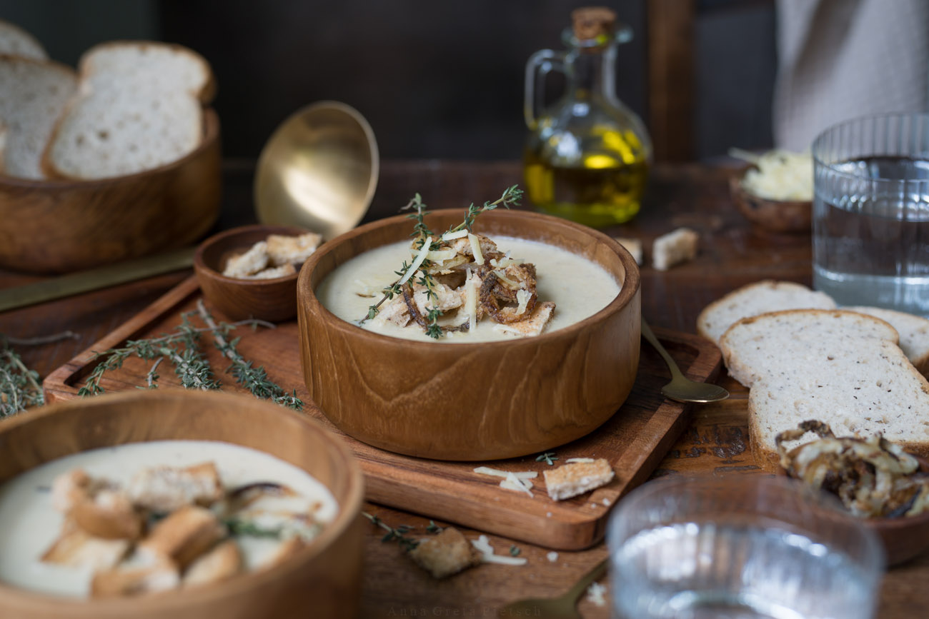 Man sieht einen Esstisch aus dunklem Holz, Darauf steht eine Holzschüssel mit einer hellen Suppe. Drumherum stehen weitere Schälen mit Suppe und Brot.