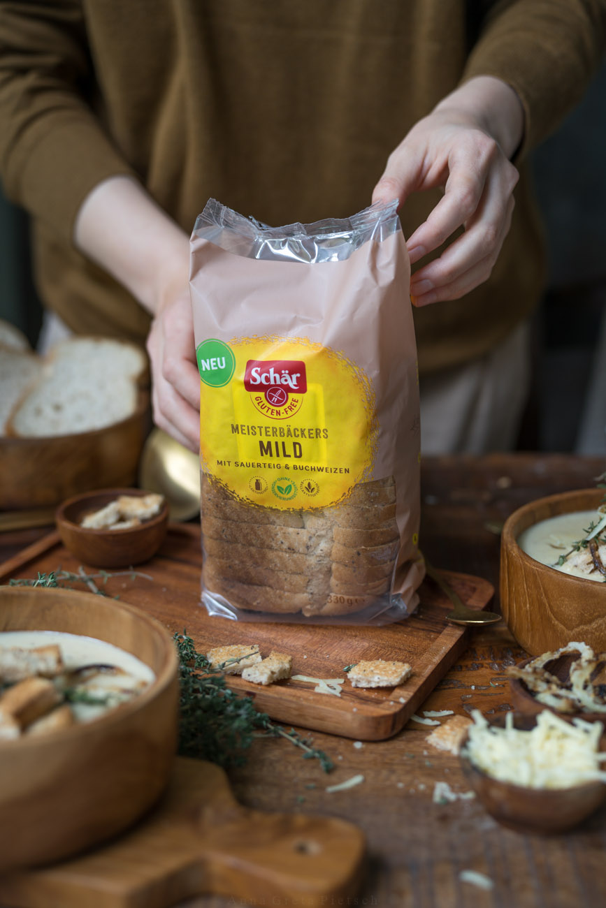 Man sieht einen Esstisch aus dunklem Holz. In der Mitte hält eine Person eine Verpackung glutenfreies Brot in die Kamera. Drumherum stehen Schälchen mit Suppe und Brot.