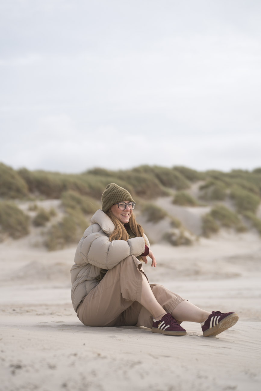 Eien Person in beigen/hellgrauen Sachen sitzt am Strand.