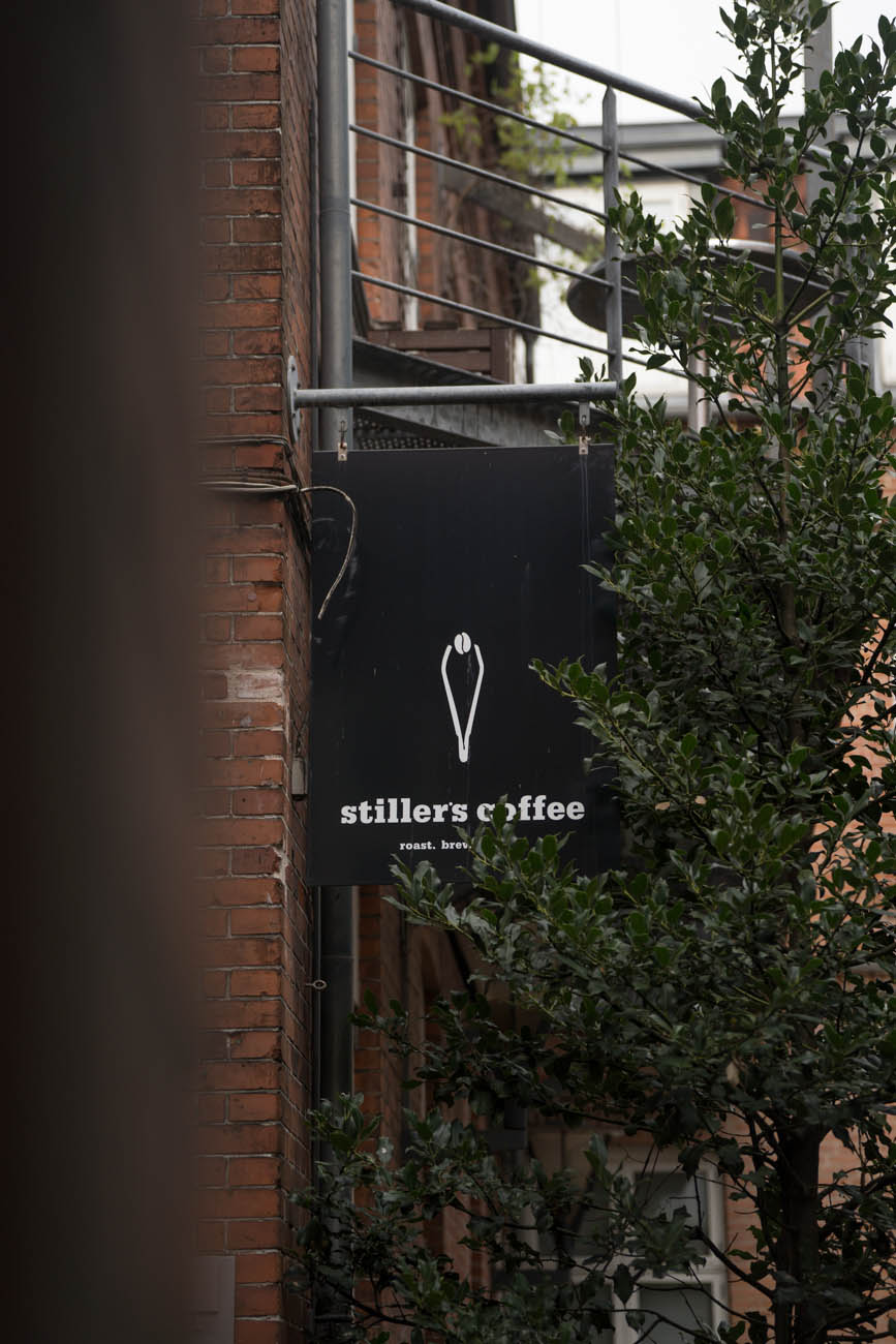 Ein schwarzes Schild mit weißer Schrift an einer Hausfassade mit dem Namen des Cafés "Stillers Coffee".