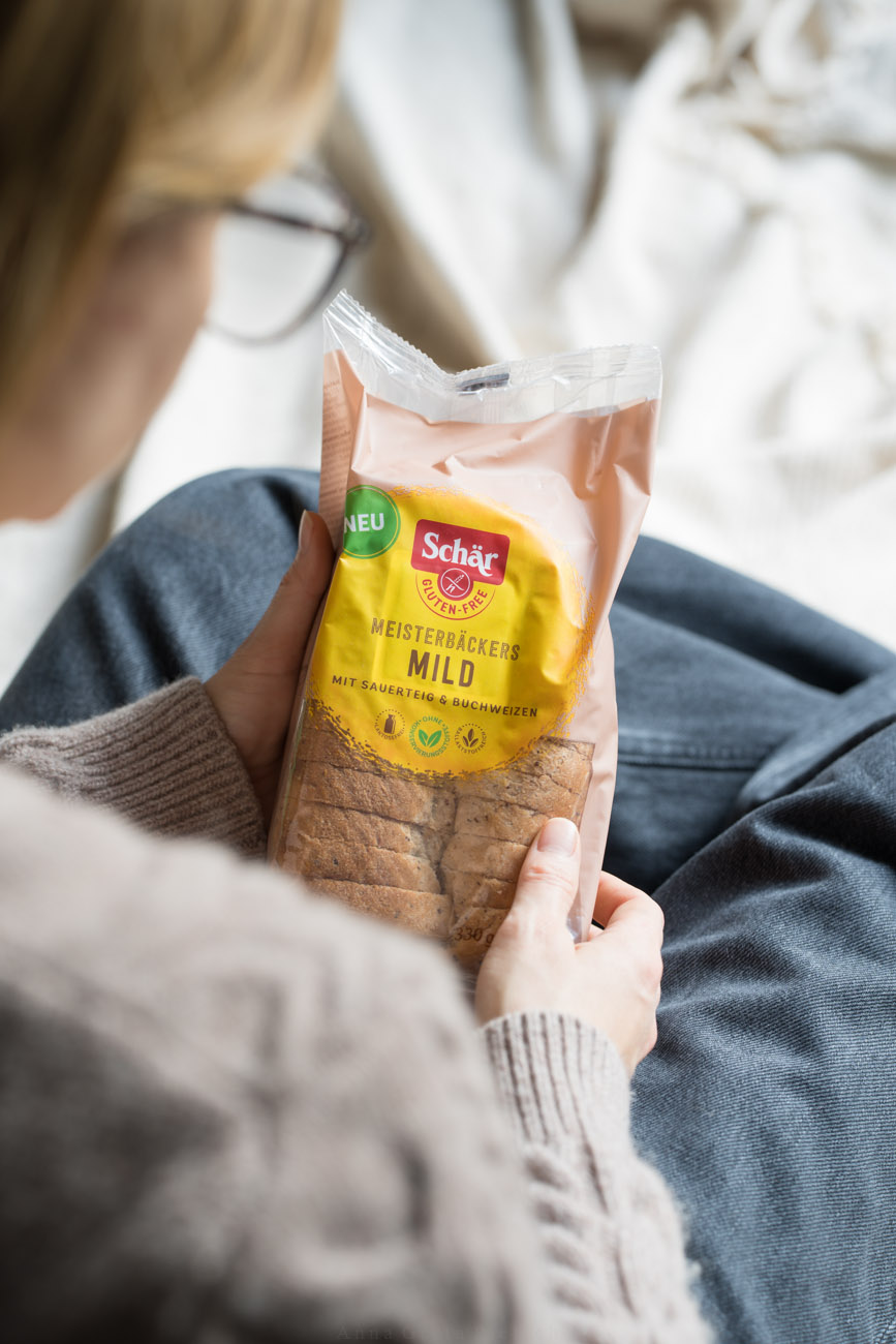 Eine Person sitzt auf einer Decke im Wohnzimmer und hält eine Packung mit glutenfreiem Brot in ihren Händen.