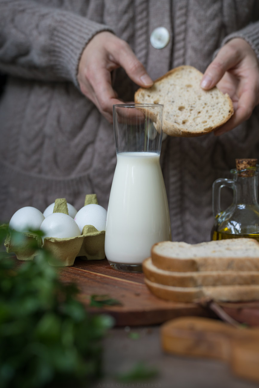 Eine Glaskaraffe mit Milch steht in der Mitte des Bildes. Darum sieht man noch gestapelte Brotscheiben und Eier im Hintergrund. Eine Person hält eine Brotscheibe in den Händen. Es sieht aus wie Zutaten für ein Rezept.