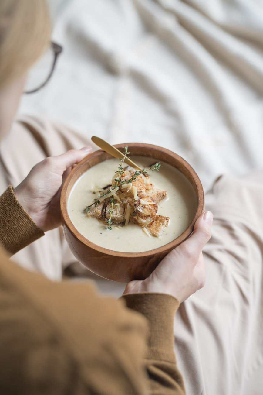 Eine Person hält eine Holzschale mit Suppe in den Händen. Das Foto wurde über die Schulter aufgenommen.