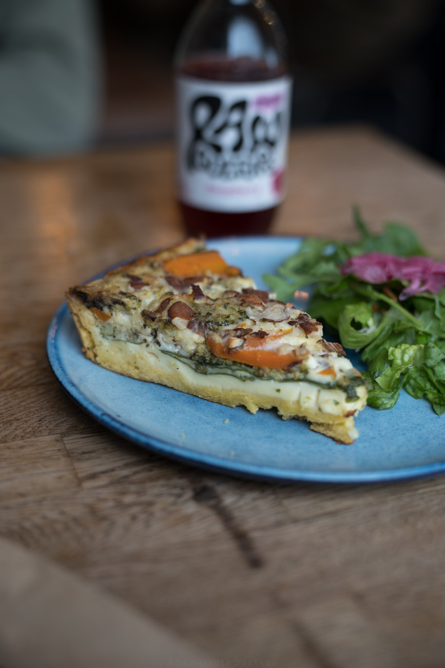 Ein Holztisch, darauf steht ein hellblauer Teller mit einem Stück Quiche. Dahinter steht eien Glasflasche mit einem Getränk.