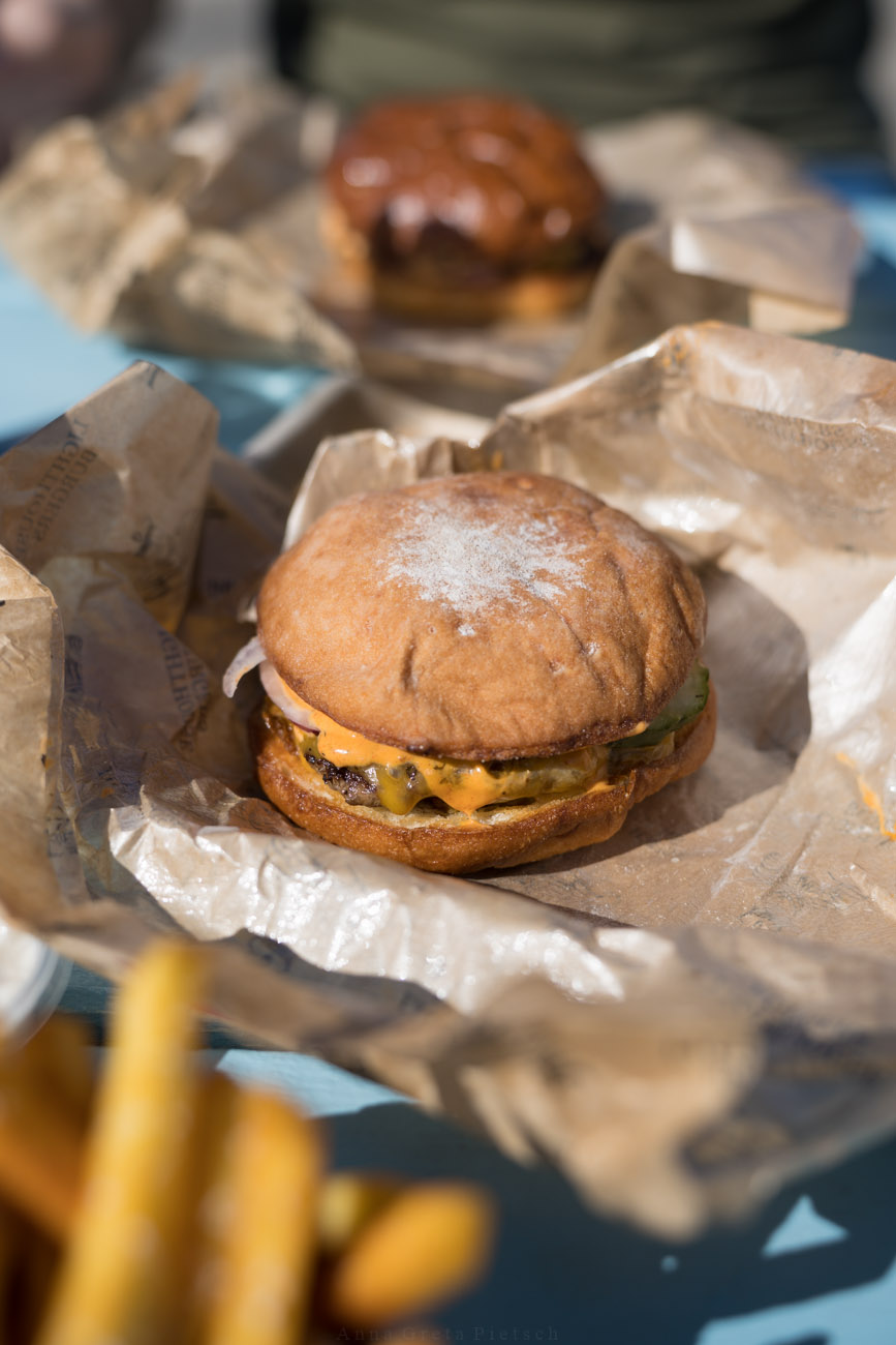 Ein Burger liegt auf einem braunen Papier auf einem Tisch. Im Hintergrund sieht man einen weiteren Burger und im Vordergrund Pommes.